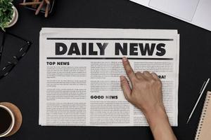 Hand pointing Business Newspaper and office supplies with notebook, hot coffee cup, glasses, books and accessories on black desk topview, Daily Newspaper mockup concept photo