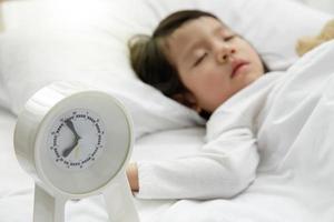 niña durmiendo con despertador en la cama de madera de su dormitorio, niña asiática feliz y despertador se despiertan por la mañana foto