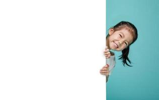 niño lindo y sonriente escondido detrás de una pizarra en blanco, espacio vacío en una foto de estudio aislada en un fondo azul colorido