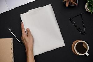 Hand holding Blank Business Newspaper and office supplies with notebook, hot coffee cup, glasses, books and accessories on black desk topview, Daily Newspaper mockup concept photo