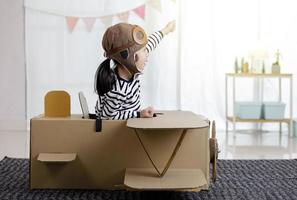 Asian little child girl playing with cardboard toy airplane handicraft in living room with copy space for your text, Creative at home and dreams of flight concept photo