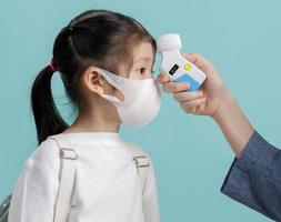 Mom and Asian little child girl wearing respirator mask to protect coronavirus outbreak and body temperature checked amid, New virus Covid-19 from Wuhan China concept, isolated on blue background photo