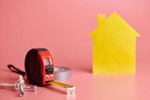Metal tape measure funny concept. House renovation. Home repair and redecorated concept. Yellow house shaped figure on pink background. photo