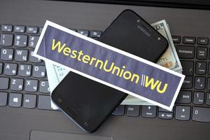 TERNOPIL, UKRAINE - SEPTEMBER 6, 2022 Western Union paper logotype lies on black laptop with US dollar bills. Western Union Company is American multinational financial services company photo