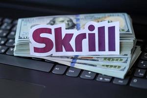 TERNOPIL, UKRAINE - SEPTEMBER 6, 2022 Skrill paper logotype lies on black laptop with US dollar bills. Payoneer is American financial services company provides online money transfer photo