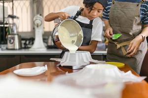 Mother with young son wearing apron pouring cream from mixing bowl into baking mold. photo