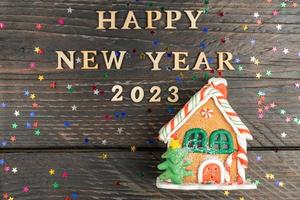 feliz año nuevo 2023 texto de madera y casa decorativa de pan de jengibre sobre un fondo oscuro con estrellas de confeti dispersas. tarjeta de felicitación festiva de navidad o año nuevo foto