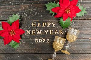 happy new year 2023 inscription made of wooden letter, red poinsettia flowers and champagne glasses on wooden background. Festive greeting card for winter holidays photo