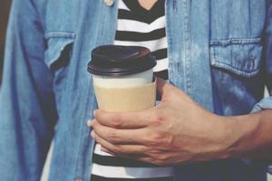 mano de hombre joven sosteniendo una taza de papel para llevar bebiendo café caliente en la cafetería. foto