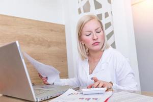 Serious focused businesswoman with laptop holding papers preparing report analyzing work results, female executive doing paperwork at workplace using computer online software for data analysis photo