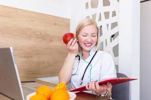Smiling nutritionist in her office, she is holding a fruit and showing healthy vegetables and fruits, healthcare and diet concept. Female nutritionist with fruits working at her desk photo