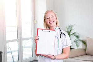 Female doctor holding clip board and smiling. Woman doctor standing straight in home care hospital. Close-up of a female doctor using clip board with stethoscope around her neck photo