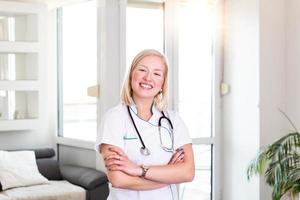 seguro médico de pie con los brazos cruzados. mujer joven sonriente segura de sí misma doctora. médico interno joven mujer bonita en ropa blanca con un estetoscopio posando y sonriendo. foto