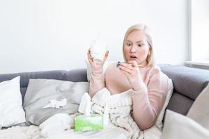 Woman is having flu and she is using thermometer. Sick with a rhinitis woman dripping nose. Woman being sick having flu lying on sofa looking at temperature on thermometer, high fever. photo