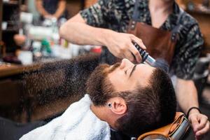 Bearded man with long beard, brutal, caucasian hipster with moustache, with stylish hair, haircut, getting powder on skin with makeup brush by barber or hairdresser hands at barbershop photo