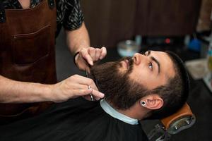 Bearded male sitting in an armchair in a barber shop while hairdresser trip his beard with scissors. Close-up of barber shearing beard to man in barbershop photo
