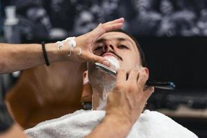 Man mith mustaches having a shave. Razor in hands of specialist barber. Young man getting an old-fashioned shave at the barber shop. Closeup, retro styled imagery photo