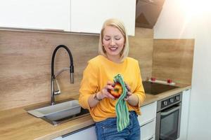 linda chica con cabello rojo rubio azotando su manzana con una toalla de cocina. mujer joven limpiando su fruta en su cocina. ingesta diaria de vitaminas con frutas, dieta y alimentación saludable foto