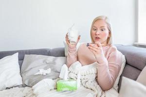 Sickness, seasonal virus problem concept. Woman being sick having flu lying on sofa looking at temperature on thermometer. Sick woman lying in bed with high fever. Cold flu and migraine. photo