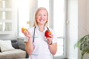 Smiling nutritionist in her office, she is holding a fruit and showing healthy vegetables and fruits, healthcare and diet concept. Female nutritionist with fruits photo