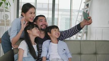 Happy Asian Thai parents, and children online video call with family via internet on sofa in home living room, lovely weekend, and domestic wellbeing lifestyle with internet technology mobile phone.