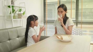 una familia tailandesa asiática saludable, una hija pequeña y una madre joven beben leche blanca fresca en un vaso y pan de alegría juntos en una mesa de comedor por la mañana, bienestar nutrición hogar desayuno comida estilo de vida. video