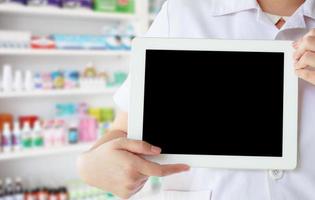 farmacéutico con bata blanca que muestra una tableta digital en blanco con estantes de medicamentos en la farmacia foto