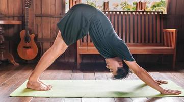 el hombre asiático deportivo de negro está haciendo yoga mientras hace ejercicio en una colchoneta de yoga en casa con piso de madera. práctica de yoga hombre pose entrenamiento concepto de vida saludable