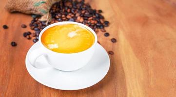 Close-up of a cup of latte with golden foam and mixed or blend coffee beans in a brown sack on an old wooden floor, top view. photo