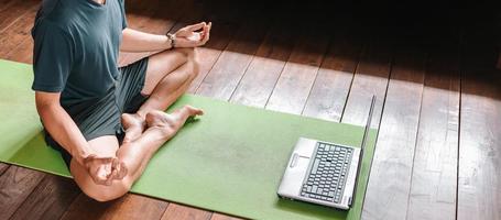 young Asian man  coach internet video online training yoga instructor modern laptop screen meditate Sukhasana posture relax breathe easy seat pose gym healthy lifestyle at home concept. photo