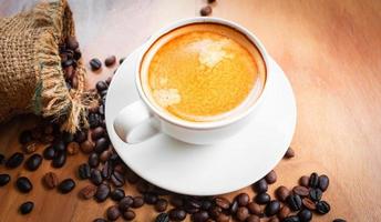 Close-up of a cup of latte with golden foam and mixed or blend coffee beans in a brown sack on an old wooden floor, top view. photo