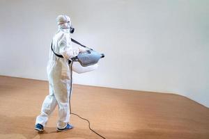 Professional technical man in prevention suit sprays sterilising solution by electrical spray machine on wood floor and white background with studio light. photo