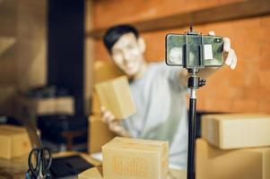 un hombre asiático usa un teléfono móvil para tomar pedidos y mostrar artículos en una caja que graba video en vivo en línea en la tienda. concepto de entrega de mercado en línea asiático propietario de pequeña empresa foto