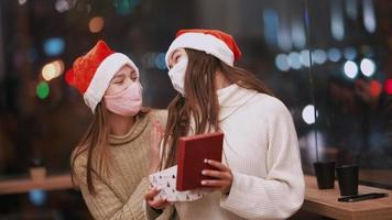 twee jong Dames in de kerstman hoeden uitwisseling cadeaus met glimlacht en knuffels terwijl vervelend maskers video