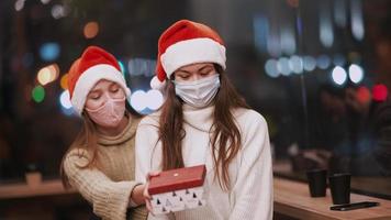Frau schleicht sich hinter Freund, um ein Überraschungsgeschenk zu enthüllen, während beide nachts in einem Café Weihnachtsmützen tragen video