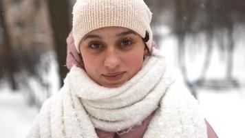 Young woman wrapped in a scarf and hat looks at camera as snow falls video
