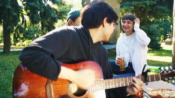 Friends sit on a blanket in the park with drinks while young man plays acoustic guitar video