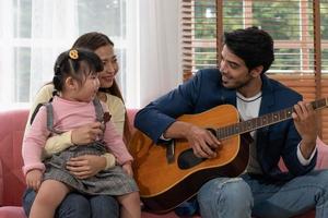 la madre asiática y el niño adoptado escuchan la canción que el padre caucásico toca la guitarra y canta en casa. concepto de familia de raza mixta juntos foto