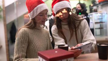 dos mujeres con sombreros de santa miran el teléfono inteligente juntos vistos a través de la ventana de un café por la noche video