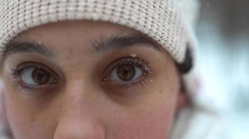 Young woman wrapped in a scarf and hat looks at camera as snow falls on eyelashes video