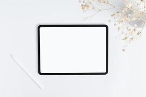 white tablet and stylus with copy space blank screen, light background with dry flower blossoms in vase, top view photo