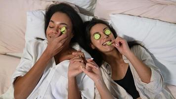Girl friends lay on bed with cucumber slices on eyes laughing and eating the cucumber video