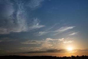 The sun goes down on the horizon. The sky is blue with white clouds. The sky glows red on the horizon. The outlines of trees can be seen at the bottom. photo