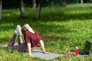 Young girl having online workout outdoors using laptop. Pilates or yoga video lesson on internet. Happy smiling girl practicing pilates lesson online in garden outdoors during quarantine.