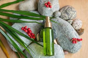 A bottle of green color with a spray bottle lies on the stones, next to the green leaves and berries of schisandra chinensis