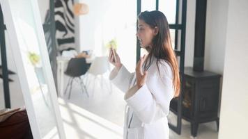 Young woman in robe video chats on smart phone standing before a mirror