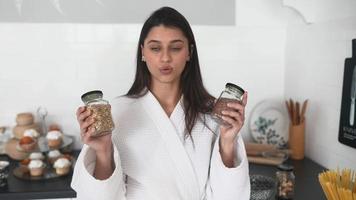 Young woman in robe holds jars of seasoning or food in bright white kitchen walks and talks toward camera video