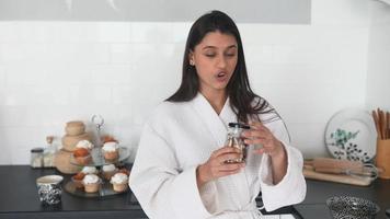 Young woman in robe holds jars of seasoning or food in bright white kitchen walks and talks toward camera video