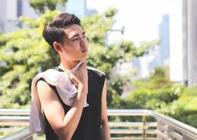 Asian young man wearing sportswear, standing and wiping sweat with towel outdoor on walkway with city building background. photo