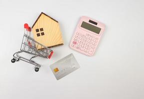 flat layout of wooden house model in shopping trolley, pink calculator and credit card on white background, home purchase calculation concept. photo